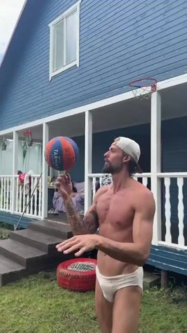 A man with tattoos on his arm and chest, wearing a white baseball cap and white briefs, stands outside a blue house with a porch. He is spinning a basketball on his index finger. A basketball hoop is mounted on the house's porch. In the background, two people are seated on a sofa or chairs. A red tire is on the grass in front of the man. The scene is outdoors, with grass and trees visible.
