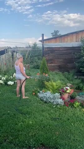A woman with blonde hair, wearing a pink tank top and grey shorts, stands in a grassy yard holding a black hose. She is watering a variety of plants, including flowers and a small evergreen tree, in front of a rustic wooden building. The building appears to be a shed or a small cabin. The sky is partly cloudy with a blue hue. The scene suggests a domestic or gardening setting during daylight hours.