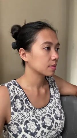A medium shot presents a woman in a sleeveless, patterned top, her dark hair pulled up in a bun and secured with a black clip. She is seated, with her left arm resting on a grey chair back. Her facial expression is thoughtful as she appears to be listening to someone or something off-camera, with her gaze directed to the right side of the frame. The background is a plain, beige wall.