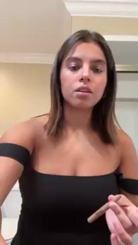 A light-skinned woman with brown hair and wearing a black, off-the-shoulder dress is applying lip liner in a bathroom. She holds a gold-colored lip liner pencil and carefully outlines her upper and lower lips, looking directly at the camera. The bathroom has beige walls and a white countertop with white tiled accents.