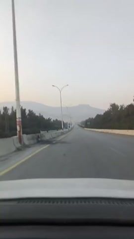 From the perspective of the driver of a white vehicle, a highway is seen stretching into the distance. The sky is overcast. The road is mostly empty, except for a few trucks pulled off to the side on the left-hand side of the road. There are trees lining both sides of the road. In the distance, there are mountains. Street lights line the road as well. The driver is moving down the road.