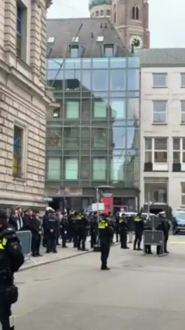 The video shows a street scene in what appears to be a European city, possibly Munich, Germany, given the architectural style and presence of the Frauenkirche (Cathedral of Our Lady) in the background. A group of uniformed police officers stands in formation in the street, seemingly maintaining order. Some civilians are standing on the sidewalk behind a metal barricade to the left of the officers. Above them, a helicopter is visible in the overcast sky. The street is paved, and the buildings surrounding the scene vary in architectural style, including a modern glass-fronted structure and more traditional stone buildings. The overall atmosphere suggests a planned or expected event requiring a police presence.
