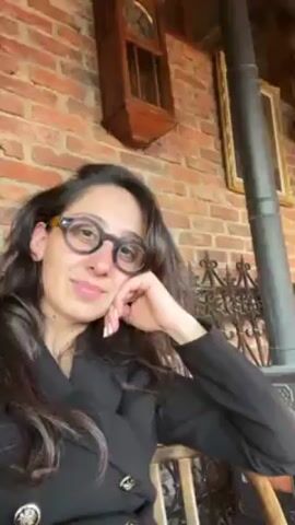 The video shows a blurred view of a woman in glasses and a black blazer sitting inside a room. The room has a wood-burning stove with a flue pipe, a brick wall, wooden paneling, a wooden shelf, a framed mirror, and an antique-style chair. The stove appears to be lit, casting a warm glow. The camera pans slightly to reveal more of the interior. The scene conveys a rustic, cozy, and warm atmosphere.