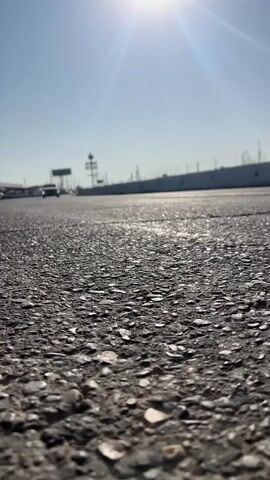 This is a low-angle, close-up shot of asphalt pavement. The sun is bright and high in the sky, creating lens flare. In the distance, a car is driving away from the camera, and beyond that, there's a white barrier and a clear blue sky. The scene appears to be outdoors on a sunny day, likely on a road or highway.