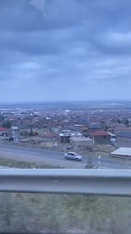 The video shows a panoramic view of a town or city from a moving vehicle, likely a bus or car, as indicated by the presence of a metal railing in the foreground and a window reflection. The sky is overcast with dark clouds. In the mid-ground, a highway or road runs horizontally across the frame, with a few vehicles visible. A cluster of houses with red roofs is visible on a hillside to the left of the road. The town itself appears to be spread out across a wide valley, with buildings stretching towards the horizon. The overall impression is one of a suburban or semi-rural landscape under a cloudy sky.
