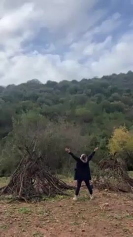 A woman stands with her arms outstretched in a clearing. She wears a dark jacket, dark pants, and light-colored shoes. Behind her is a hillside covered with trees and a cloudy sky. To either side of her are piles of dead branches, formed into teepee shapes. The ground is a reddish-brown dirt and scattered with small plants.