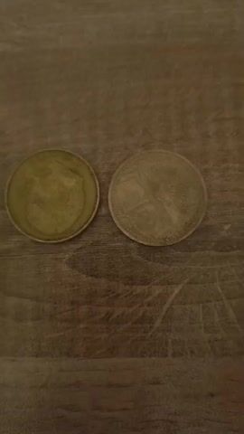 The video shows a close-up of two coins lying on a wooden surface. A hand enters the frame and manipulates the coins. One coin appears to be gold-colored with an image on it, while the other is silver-colored and blank on the visible side. The hand briefly covers the silver coin and then moves away, leaving both coins visible again.