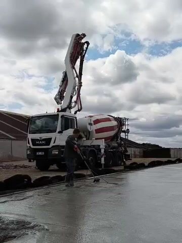 The video shows a construction scene with a concrete mixer truck and a man working on a concrete surface. The truck, a white MAN model with a red and white striped concrete drum, is positioned on the left side of the frame. Its boom is extended over a newly poured concrete slab. A man, wearing a dark jacket, jeans, and boots, is standing on the wet concrete and using a long-handled tool, possibly a rake or broom, to smooth the surface. The environment appears to be outdoors, with a cloudy sky overhead and some buildings visible in the background. The concrete slab is dark and wet, indicating it has been recently poured. The man's actions suggest he is in the process of finishing or leveling the concrete.