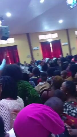 This video captures a vibrant indoor gathering inside a large hall, likely a church or event center, in Ogoja, Nigeria. The setting features a high, light-colored ceiling equipped with numerous recessed lights and hanging speaker units. The walls are also light-colored, accented by prominent red curtains covering windows or doorways. A large screen is visible near the ceiling in the initial frames.

The hall is densely packed with a diverse crowd of people, including many adults and several young children, some of whom are being held. Most attendees are standing and appear to be actively participating in an event. Many individuals are seen raising their hands, suggesting worship, celebration, or enthusiastic engagement with a speaker or music. People are swaying and moving with energy, contributing to a lively atmosphere. Clothing worn by the attendees is varied, with some individuals in bright colors such as pink, green, and blue, as well as patterned outfits. The camera is handheld, moving through the crowd, offering a dynamic perspective of the gathering. The audio contains speech, including a mention of "August" and a call to "join up," indicating an organized event or service. No discernible text is visible within the video frames.