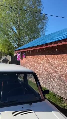 The video shows an outdoor scene on a bright, sunny day. In the foreground, a white car with its hood visible is parked. Behind it, there is a building constructed from OSB (Oriented Strand Board) with a blue metal roof. Two "OZON" signs, a Russian e-commerce company, are attached to the building. A string of lights is strung below the roofline. As the camera pans right, the full extent of the building becomes visible, which extends into another building constructed from similar material with a darker red-brown hue. A chain-link fence runs along the front. A person wearing a red top and sunglasses exits the building, while another person is seen in a window. The background features green trees and a clear blue sky.