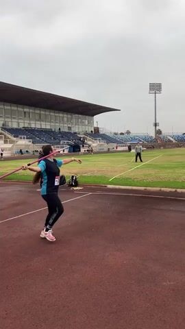 A woman is practicing javelin at a stadium. She is wearing athletic clothing and holding a javelin. The stadium has a track, a field, and bleachers. The sky is cloudy.