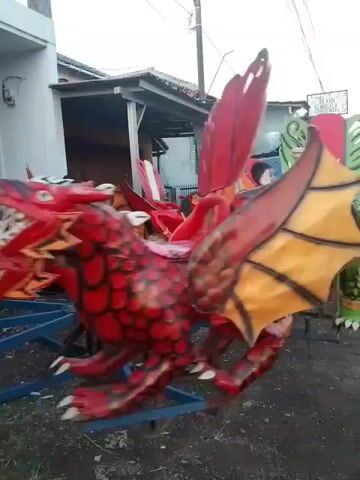 The video shows a children's ride with a carousel-like design, featuring various dragon and monster-shaped seats painted in vibrant colors such as red, blue, green, and pink. Children are riding on the attraction. The ride is set up outdoors in what appears to be a residential area, with houses and a utility pole visible in the background. The ground is a mix of dirt and asphalt. The ride is rotating.
