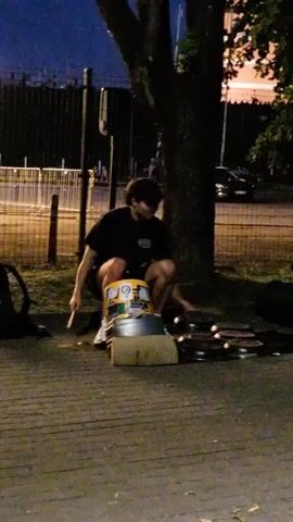 A person wearing a black t-shirt and dark shorts is crouched down on a paved surface next to a tree. They are holding sticks in both hands and appear to be playing a set of round, metallic objects laid out on the ground in front of them, possibly cymbals or tambourines. There is a yellow and silver bucket in front of the person. In the background, there is a metal fence, other people, and vehicles. The scene is dimly lit, suggesting it is dusk or nighttime.