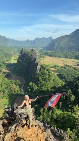The video shows a woman standing on a rocky outcrop, holding a flag on a pole. She is wearing a black sports bra and green pants. The flag is red and white with some black. She is looking out at a vast landscape of green hills, valleys, and mountains. The sky is blue with some clouds. The sun is shining. The woman is in the foreground, and the landscape is in the background. The video is shot from a high angle, looking down at the woman and the landscape. There is a bicycle to the left of the woman. The woman is waving the flag. The video is static.