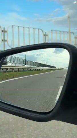 The video shows a view through a car's side mirror. The mirror reflects a road with a white dividing line and a barrier on the right side. The sky is visible with some clouds. A large metal structure, possibly a bridge support or tower, is visible in the distance. The perspective suggests the car is moving on a road.