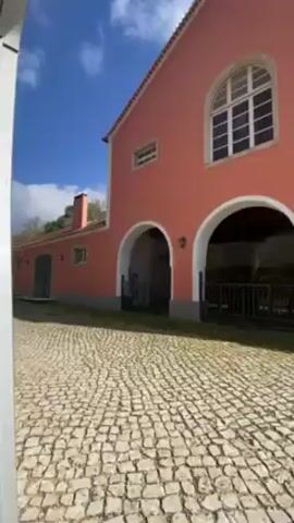 The camera moves forward showing a cobblestone courtyard in front of a two-story, salmon-colored building with a red-tiled roof. The building has three arched openings with wrought iron gates and a large arched window above. To the left is an extension of the building with a chimney and a dark green door. Small decorative lamps flank the arches. The sky is bright blue with scattered white clouds.