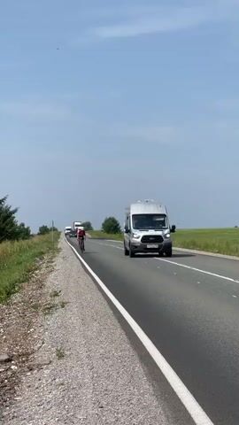 A cyclist wearing a red and white jersey and a white helmet is riding a bicycle on the right side of a paved road. The cyclist is moving away from the camera, with a white line marking the edge of the road to their left. A silver van is driving in the same direction as the cyclist, on the left side of the road and is passing the cyclist. In the distance, there are other vehicles, including a truck. The road is surrounded by a grassy field on the right and a gravel shoulder on the left. The sky is clear and blue with some clouds. The overall scene depicts a cycling race or training session on a rural road.
