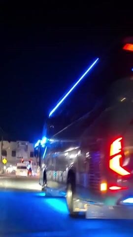 A nighttime exterior shot captures the rear of a large bus as it drives away from the camera. The bus is illuminated with numerous colored lights, including blue lights along its top edge and around its taillights, and red and pink lights reflecting off the wet road surface beneath it. The bus has several decorative lights on its rear, including what appear to be stylized red eyes and text that is difficult to read due to the lighting and resolution. The bus is on a paved road with a sidewalk and buildings visible on the left side of the frame. The sky is dark, indicating it is nighttime. Other vehicles with headlights are visible in the distance.