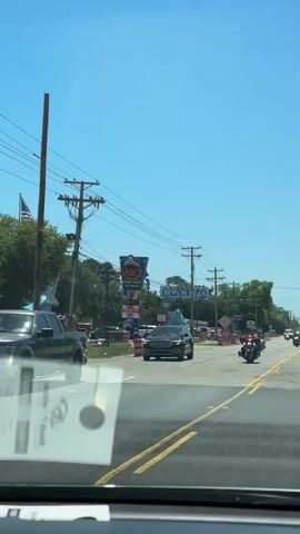 The video is taken from inside a car looking out the windshield at a street with moderate traffic. The sky is clear and blue. On the left side of the road is an American flag, power lines, and trees. There is a shark-themed business sign. On the right side of the road is a sign that reads "SEVEN SEAS." Traffic, including a line of motorcycles, is visible. The video is steady. The lighting is bright.