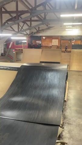 A person on a scooter is in an indoor skatepark. The person is wearing a white shirt, dark shorts, knee pads, and a helmet. The person is riding down a ramp made of dark material. In the background, there are other ramps and structures made of wood. The skatepark has a high ceiling with exposed beams and industrial lighting. The camera is mounted low and moves with the scooter rider.