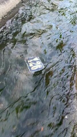 The video shows a concrete canal or drainage ditch with clear water flowing through it. In the foreground, a white square object with the number "126" written in dark blue or black is floating on the surface of the water, being carried along by the current. The camera pans upwards, revealing the length of the canal which is lined by concrete banks. Green trees are visible along both sides of the canal in the background, suggesting an outdoor setting. On the left side, further in the distance, a concrete or stone wall is visible, and a car can be briefly seen. The scene depicts a calm, flowing waterway on what appears to be a clear day in Târgu Mureș, Romania.