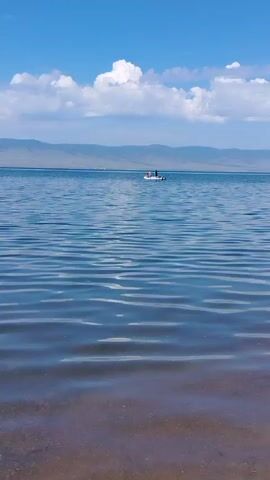 The video shows a serene lake with gentle waves under a bright blue sky dotted with white clouds. In the distance, a small boat with several people on board is visible. The shore in the foreground is covered with pebbles. A fishing rod is partially visible on the right side of the frame. The overall scene evokes a peaceful outdoor activity on a clear day.