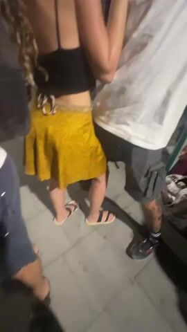 A couple is dancing outdoors at night. The woman is wearing a yellow skirt and the man is wearing a white t-shirt. The camera is shaky and focuses on the couple's faces as they dance and interact. The background is dark with some blurred lights and structures.