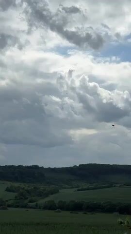 The video shows a panoramic view of a valley with rolling green hills and a dramatic cloudy sky. The landscape is dominated by lush vegetation, with patches of darker green suggesting forests and lighter green areas likely representing fields. A distant treeline forms a dark, undulating horizon. The sky is filled with a dynamic display of clouds, ranging from dark, heavy formations to lighter, wispy ones. Sunlight occasionally breaks through, casting illuminated patches on the landscape. A small, dark object, possibly a bird or an aircraft, is visible in the sky towards the upper right quadrant of the frame. The overall impression is one of a vast, natural landscape under a constantly changing sky.
