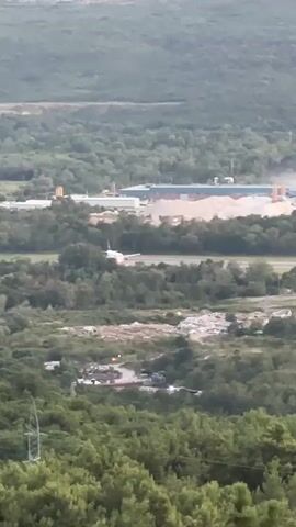 A long-shot video shows an aerial view of a landscape with trees, a building, a construction site, and an airplane on a runway. The video is from the perspective of someone looking down on the scene from a higher elevation. The airplane is white with a red tail and is on a runway that appears to be part of an airport. There is a large pile of sand or dirt next to the runway and a building in the background that appears to be a factory or warehouse. The sky is overcast and the lighting is dim, suggesting it is either early morning or late evening.