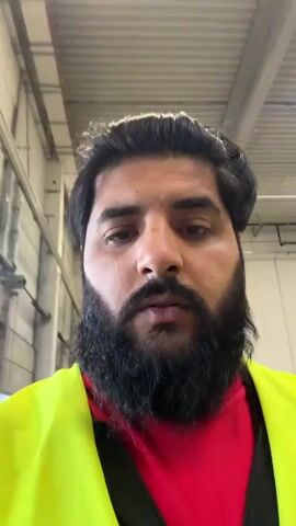The video shows a close-up, head-and-shoulders shot of a man with dark hair and a full beard. He is wearing a bright yellow safety vest over a red shirt. The man appears to be indoors, possibly in a warehouse or industrial setting, as evidenced by the high ceiling with exposed beams and lighting fixtures, and the metallic structure visible in the background. He is looking directly at the camera and appears to be speaking, though no audio is provided. The lighting is bright, suggesting artificial illumination. The camera is mostly stationary, with slight movements and slight reframing between frames.