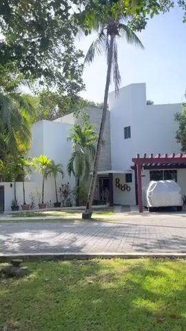 The camera is positioned near street level, facing a modern, white, two-story house in a tropical setting. The house has a carport with a dark red pergola and a car covered with a silver tarp.  The yard is green with a brick driveway and a variety of trees including several palms. The sky is bright and the sun is visible through the trees. The camera is moving slightly from left to right.