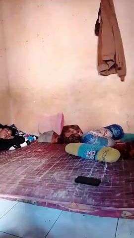 A person is sleeping on a floor covered with a patterned mat. The room appears to be dimly lit with textured walls. There are several pillows on the floor, including one with a cartoon character design. A mobile phone is placed on the mat near the pillows. A brown jacket is hanging on the wall. In another scene, a person's arm is visible. A small plate with what appears to be food is on a surface, with two glasses nearby. Further into the video, a large appliance, possibly a refrigerator, is partially visible. The scene shifts to an area with a red motorcycle, a bucket, and what seems to be a wooden cabinet.