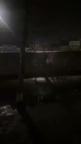 The scene is a dark, rainy night. A street lamp illuminates a wet, possibly icy ground area, with a thin tree in the foreground. Beyond the tree, there's a wall, and in the distance, stacked shipping containers are faintly visible, with the word "JIEAN" legible on one. There are no people present. The only movement comes from what appears to be rain or light snowfall.