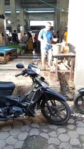 A man in a blue shirt and white pants is standing in a market. He is reaching into a bucket on a table. To his left, a black motorcycle is parked on a paved surface. In the background, other people are visible, and there are market stalls with goods on display. The market appears to be covered by a roof supported by columns.