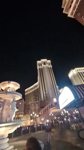 The video is shot outdoors at night, showcasing a grand view of The Venetian hotel and casino complex in Las Vegas. In the foreground, a multi-tiered fountain with classical statues is brightly lit, its water feature adding a dynamic element. The camera pans slightly to the left, revealing more of the surrounding architecture, including the Palazzo hotel, with its name illuminated on top. Streetlights line the walkways, and there is some visible traffic in the background. Despite the presence of people in the area, they are not the focus of the shot. The overall impression is that of opulence and architectural grandeur, characteristic of the Las Vegas Strip.