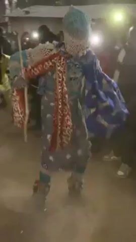 A person in an elaborate, colorful costume dances in a dirt area surrounded by people. The dancer’s costume includes a patterned blue and white hat, a patterned blue and white shirt with what appear to be wings, and a multi-colored garland. Many of the surrounding people are holding up phones with their flashlights on, illuminating the scene. Dust is kicked up as the dancer moves.