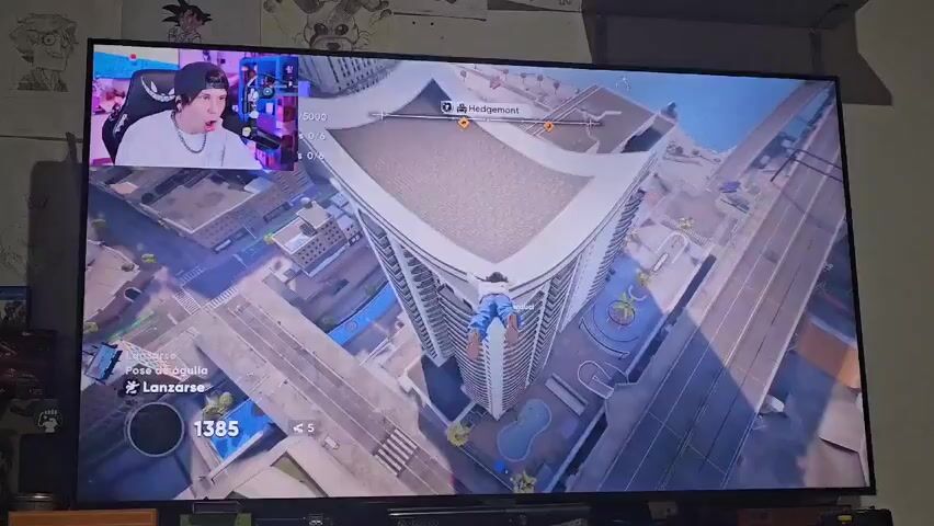 Here’s a concise description of the video:

The video shows a person playing a skateboarding video game on a large TV screen. A smaller window in the top left corner shows a streamer, presumably the person playing, reacting to the gameplay. The video game features a skater performing tricks on top of a skyscraper. The streamer appears surprised and animated in his reaction. The game displays in-game metrics and messages like "INCREÍBLE!" and "LEGENDARIO!" as the skater performs stunts. The environment around the TV includes posters and collectibles.