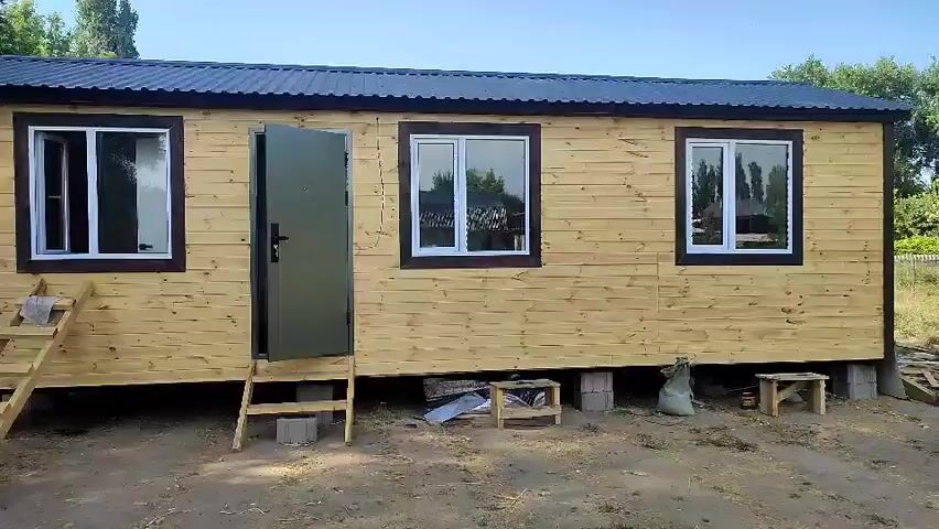The video shows the exterior and interior of a small, newly constructed wooden building. The exterior is clad in light-colored horizontal wooden planks with dark trim around the windows and roof. There are three windows and one door visible on the front of the building. The building is supported by concrete blocks. To the left of the building, there is a wooden ramp leading to the door. To the right of the building, there is a pile of lumber and some construction debris. The interior of the building is unfinished, with walls and floor made of oriented strand board (OSB). There is a white circular object on one of the walls, possibly a ventilation cover. In one shot, a watermelon and a knife are visible on the floor.