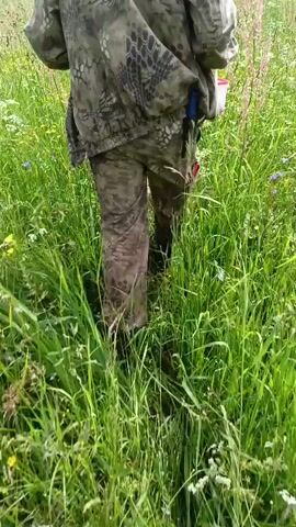 A person walks through a field of tall grass and wildflowers. The person is wearing camouflage clothing and dark boots. The field is green with patches of yellow and white wildflowers, and some taller, reddish grasses. In the distance, there is a line of trees under a cloudy sky. The camera is positioned behind the person, following them as they walk.