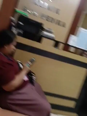 The video begins with a woman sitting in a waiting area, looking at her phone. Behind her is a counter with a sign that reads "KLINIK BUK MEDIKA". A printer and some papers are on the counter. The camera pans to the right, showing a woman sitting at a computer and a man standing behind her. Further to the right, there's a door labeled "FARMASI" and a television screen displaying an image. The camera then pans to the left again, showing two doors with signs that are difficult to read. A woman wearing a hijab and a mask walks into the frame and then towards a door labeled "R. TINDAKAN". To the right of this door, there's a security camera. Another person is visible sitting in a chair next to a scale. The video ends with a partial view of another person. The environment appears to be a clinic or hospital waiting area.
