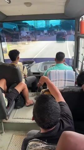 The video shows the interior of a bus moving on a road. Several passengers are visible, some facing forward and others looking out the windows. One passenger is in the driver's seat, and another is in the co-pilot's seat. The view through the windshield shows a street with buildings and other vehicles. The camera pans around the bus, capturing different angles of the passengers and the scenery outside. The overall mood appears to be casual and relaxed.