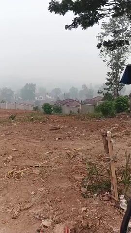 The video captures a wide shot of a hazy, elevated landscape. In the foreground, a patch of disturbed, reddish-brown earth is visible, with scattered debris and some thin yellow string or wire. A wooden stake with wire is also present. Behind this, a low, light-colored wall stretches horizontally, separating the foreground from a residential area. Beyond the wall, several houses with reddish-brown roofs are nestled amongst trees. The background is heavily obscured by fog or haze, creating a soft, blurred effect of distant trees and structures. The overall atmosphere is muted and subdued, possibly due to atmospheric conditions or time of day. The foliage of a large tree hangs into the top left corner of the frame.