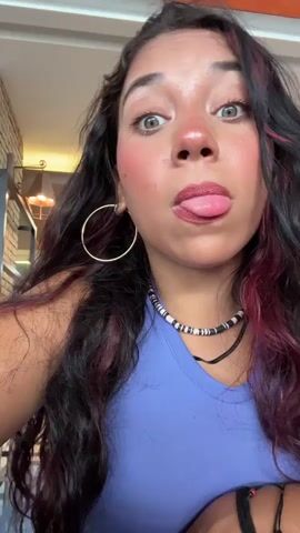The video shows a close-up, selfie-style shot of a young woman with long, dark, wavy hair with red highlights. She is wearing a purple tank top and a necklace with black and white beads. She has large hoop earrings. In the first few seconds, she sticks her tongue out, then pulls it back in and looks at the camera with a neutral expression. The background appears to be indoors, possibly a cafe or restaurant, with blurred lights and structures visible.