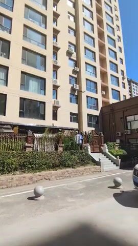 This video shows a tall, beige apartment building with many windows and air conditioning units. In front of the building is a paved area with a road and several large, gray spherical bollards. A walkway with a hedge and a decorative fence runs along the building. A set of concrete stairs leads up to an entrance, and a man is visible standing near the stairs, near a washing line. The sky is clear and blue, indicating a sunny day.