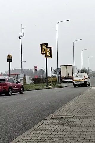 The video shows a street scene on a cloudy day with light snow falling. On the left side of the road, a red SUV is parked next to a McDonald's sign. Across the street and slightly to the right, a sign with two large "B"s is visible. Further down the road, a white truck is turning, and a van with "Garwaz" written on the side is driving away. The view is taken from a sidewalk paved with interlocking bricks. Overhead, there are streetlights and a communication tower in the background.