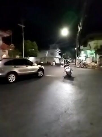 The video shows a nighttime street scene with an archway in the background. Several vehicles, including a silver SUV and motorcycles, are visible. Some people are riding on the motorcycles. The street appears to be paved, and there are trees and buildings lining the sides. The overall lighting suggests it is dark, with artificial lighting from streetlights and vehicles. The camera is moving, creating a slightly blurred effect.