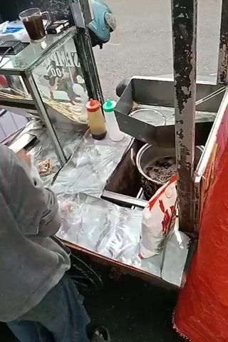 A person wearing a gray hoodie and blue jeans stands behind a food cart. The cart has a glass display case on the left, containing skewers of food. A cup of dark liquid sits on top of the display case. To the right of the display case, there are two bottles, one with a red cap and the other with a green cap. A metal basin containing boiling liquid and noodles is situated further to the right. A bag with red and white packaging sits on a surface in front of the basin. The person is reaching into the basin with a utensil, seemingly cooking or preparing food. The environment appears to be outdoors, with a paved surface visible in the background.