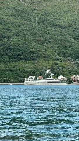 A wide shot shows a large white yacht anchored near a lush green, tree-covered hill. Several white buildings with red-tiled roofs are visible on the shore behind the yacht. The water in the foreground is a deep blue with whitecaps, indicating light waves. The overall scene suggests a tranquil coastal environment.