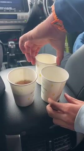 Inside a car, two paper cups sit on the center console. The cup on the left is filled with tea, while the cup on the right is empty. A person in a blue jacket is seen filling the empty cup with liquid from a green thermos. The car's dashboard and steering wheel are visible. Through the windshield, a blurry, possibly rainy or snowy, outdoor scene can be seen. There are two people in the car. The person pouring the liquid is the driver.