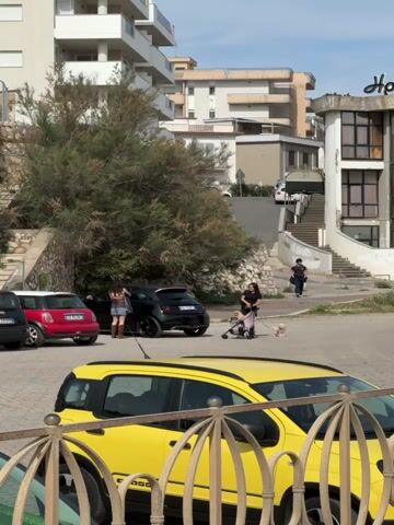 The scene shows a parking area with several parked cars, including a yellow Fiat. Two women are present: one is standing near a black car and appears to be opening the trunk. The other is pushing a stroller and walking a small white dog on a leash. A third person, possibly a man, is walking uphill on a set of stairs in the background. The surrounding area consists of buildings and greenery. The weather seems clear.